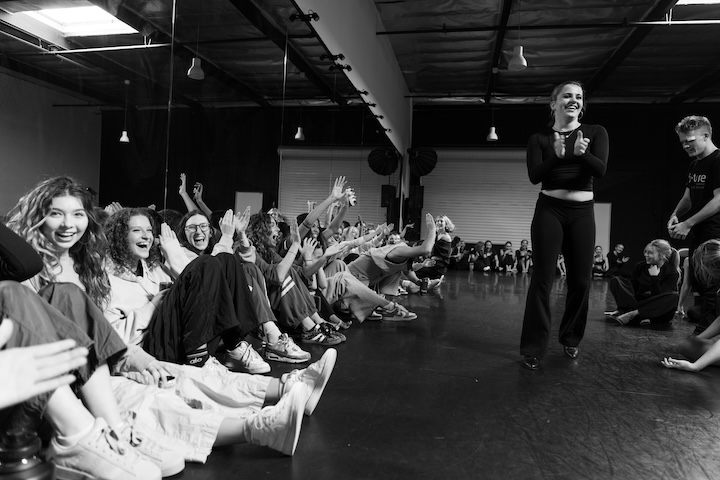 A group of diverse young dancers in a circle on a studio floor, listening intently to a mentor choreographer. The atmosphere is supportive and collaborative, capturing the community aspect of a pre-professional dance program in Orange County.
